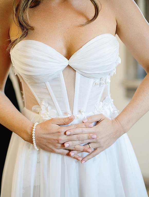 Wedding dress close-up showing strapless wedding dress corset bodice with pearl beading and floral appliqués, bride’s ringed hands at waist indoors