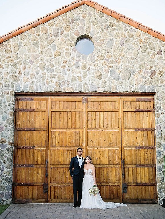 Couple portrait of bride in strapless gown holding bouquet beside groom in black tuxedo, posed by rustic barn doors and stone wall