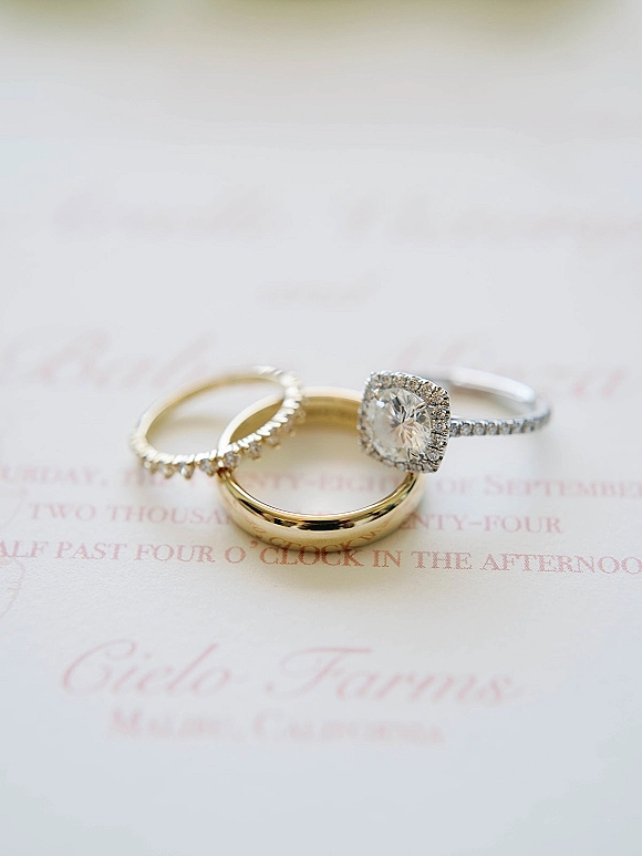 Wedding rings stacked with a diamond halo engagement ring on a blush calligraphy invitation card over white paper flat lay