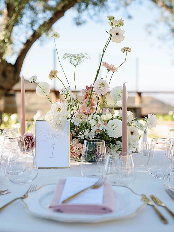 Reception tablescape with wedding table centerpiece, blush taper candles, gold flatware, and menu cards set outdoors under trees