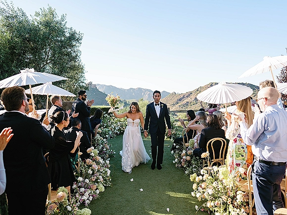 Wedding recessional as bride and groom walk the aisle, bride raising bouquet, guests under white parasols on a mountain-view lawn