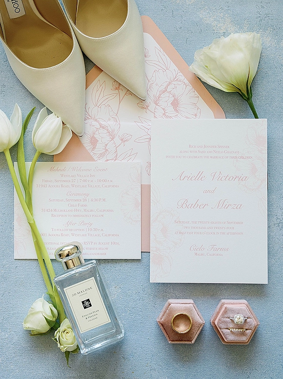 Wedding invitation suite with blush floral wedding invitations, envelope, rings in velvet boxes, heels, tulips, and perfume on blue fabric