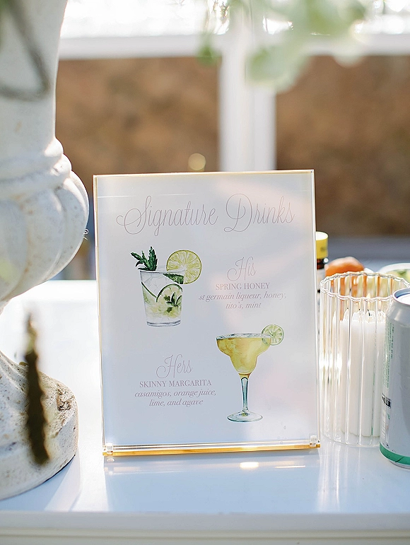 Signature drinks sign in acrylic holder with watercolor cocktail and lime garnish illustration, candles and cans on an outdoor patio bar counter