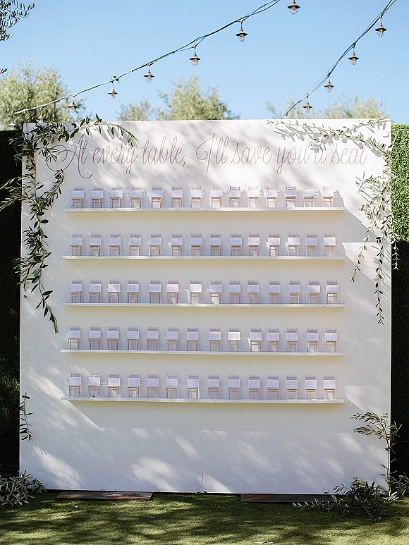 Seating chart display with escort cards on shelves, calligraphy lettering and greenery garland on a white panel backdrop with string lights outdoors