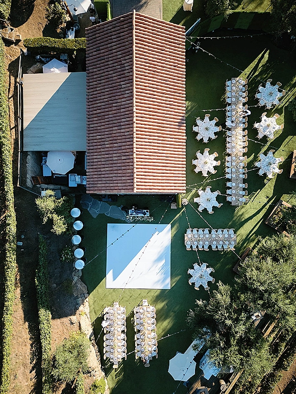 Outdoor wedding reception with aerial wedding reception layout, long and round white-linen tables, dance floor, and string lights on a lawn patio