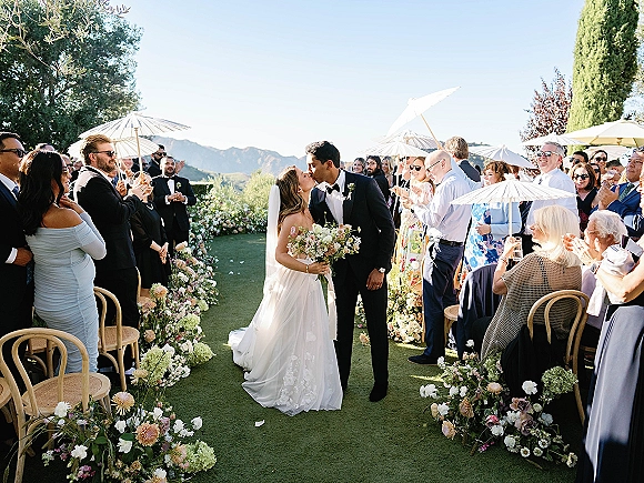 Ceremony kiss as bride in strapless wedding dress and veil kisses tuxedo groom, bouquet in hand, guests with white parasols on a mountain lawn aisle