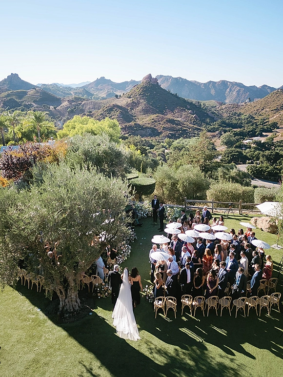 Wedding ceremony with bride in veil walking down a curved outdoor wedding ceremony aisle, guests holding white parasols, mountain view backdrop