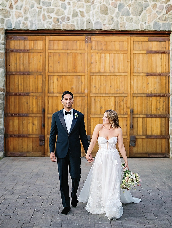 Couple portrait of bride and groom holding hands, bride gazing at him in courtyard by wooden doors, bouquet and tuxedo visible