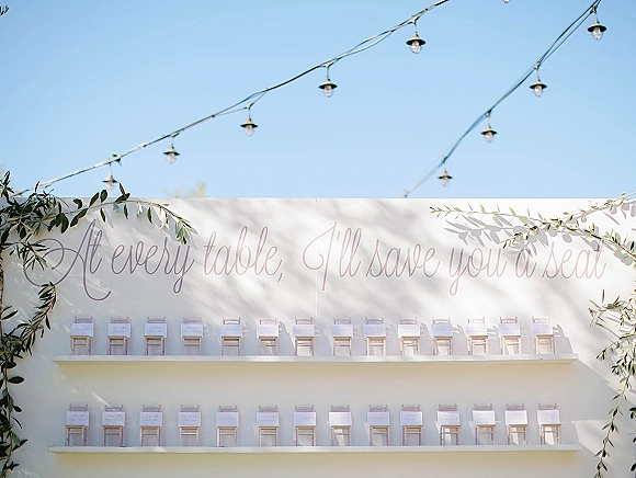 Seating chart display with wedding seating chart escort cards on shelves, calligraphy sign, greenery garland, and string lights on a white wall backdrop