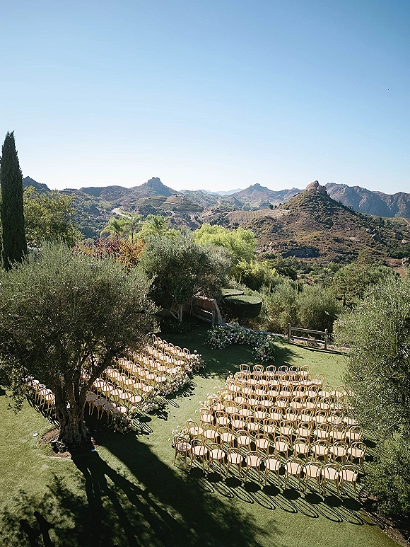Ceremony setup with outdoor ceremony seating, curved wood chairs and aisle floral arrangements on a lawn facing mountain landscape