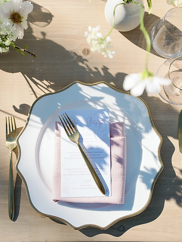 Reception place setting with a wedding menu card on a scalloped edge charger, gold flatware, linen napkin, glasses, and bud vase on a wood table