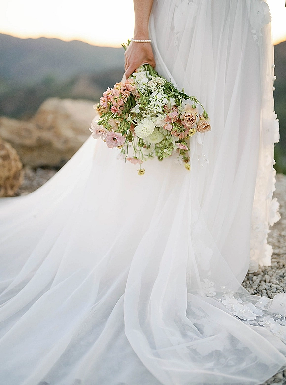 Bridal bouquet with blush roses and white flowers, held by a bride in a lace dress and pearl bracelet against rocky mountains backdrop