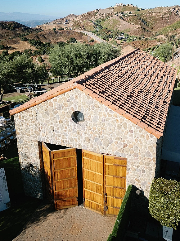 Wedding venue exterior with a stone barn, terracotta roof and open wooden barn doors, string lights over outdoor dining with mountain views