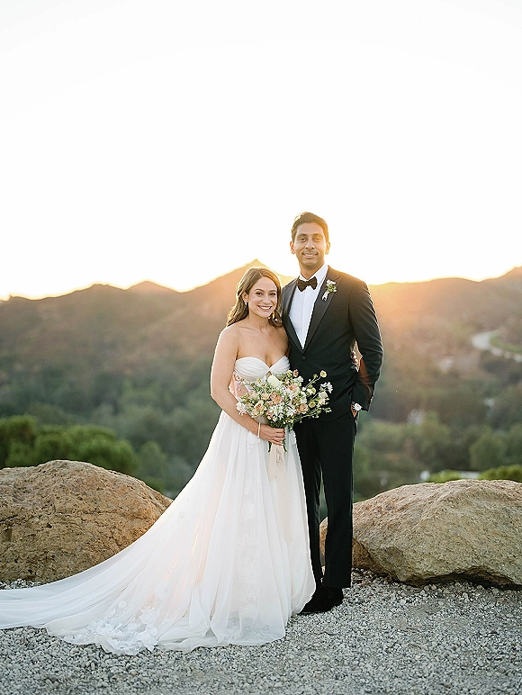 Couple portrait of bride in a strapless wedding dress holding a bouquet beside groom in tuxedo at sunset mountain overlook with boulders