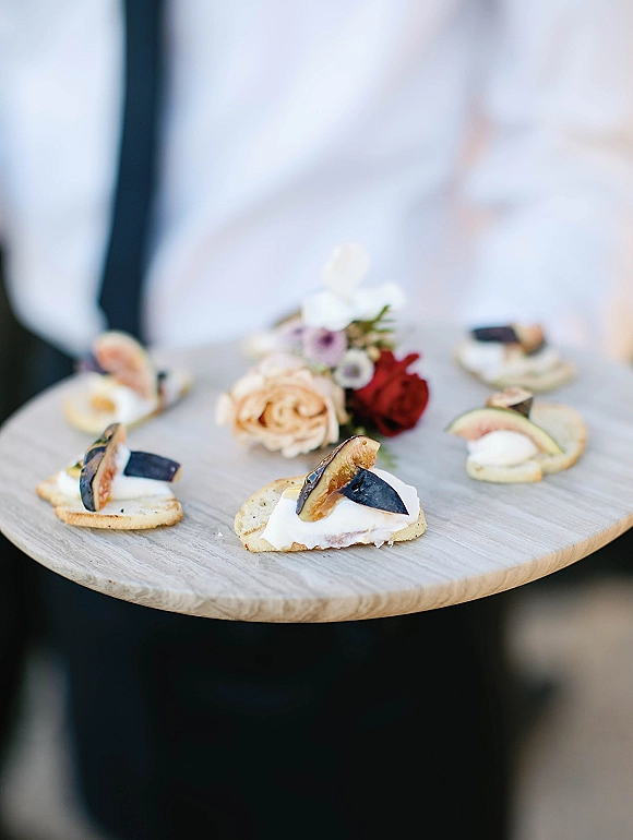 Wedding appetizers on a wooden serving board with fig and cheese crostini and edible flower garnish, held on a tray outdoors