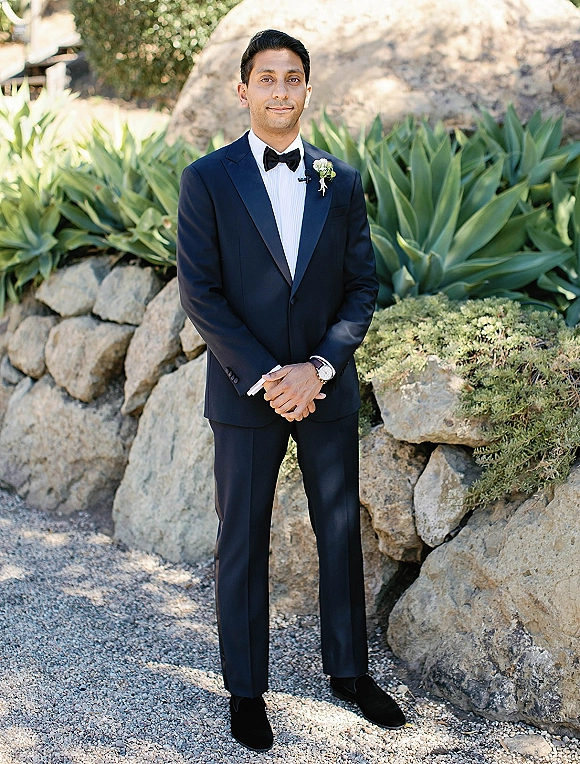 Groom portrait in a black tuxedo with bow tie and boutonniere, standing full-length on a gravel path by a stone wall and agave plants