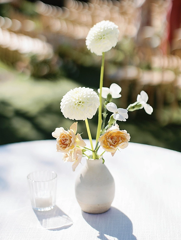 Wedding table centerpiece with a white dahlia and peach roses in a ceramic bud vase on linen, beside a ribbed glass candle holder outdoors