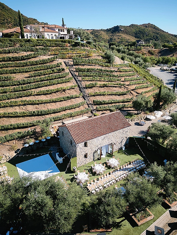 Outdoor reception setup in a vineyard wedding reception with long banquet and round tables, wooden chairs, string lights, and umbrellas by a stone building