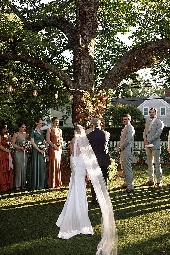 Wedding ceremony with the bride in a long veil walking toward the altar as the wedding party lines up under string lights by a large tree