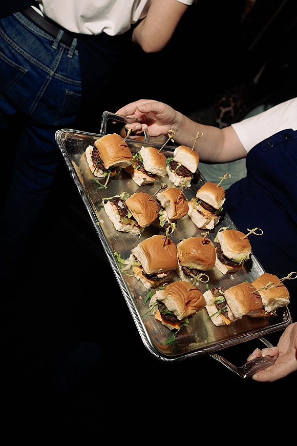 Wedding cocktail hour food of mini burger sliders on a metal tray with cocktail skewers, served indoors among reception guests