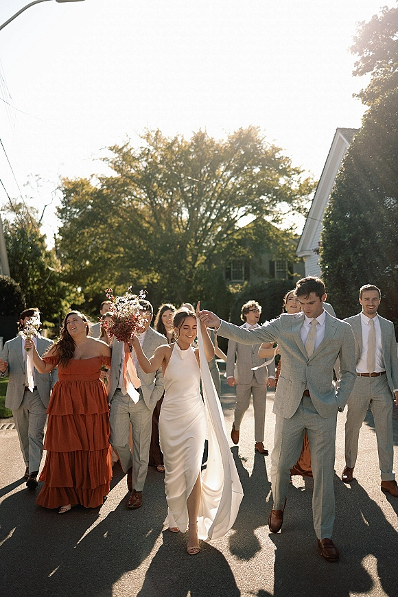 Wedding party walking with bride and groom strolling hand in hand, veil blowing, holding a wildflower bouquet on a sunlit residential street