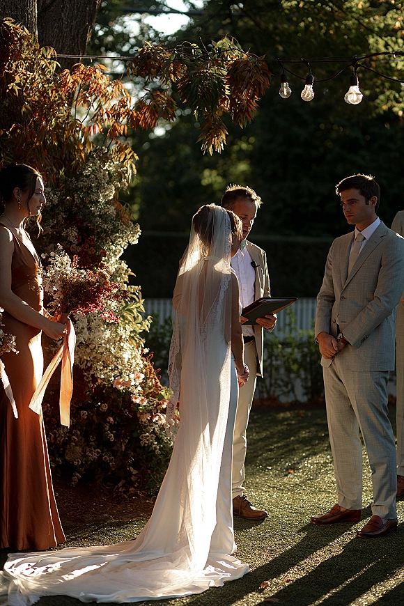 Wedding vows as bride in veil and gown train faces groom in light suit while officiant reads under floral arch in sunny garden lawn