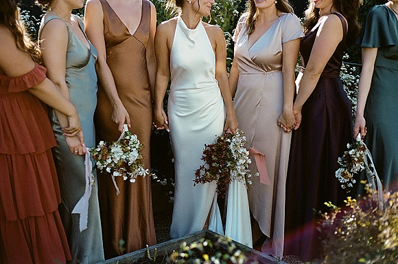 Bridesmaid group photo of bridesmaids holding hands with mismatched dresses and wildflower bouquets with ribbon streamers on a garden walkway
