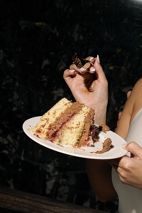Wedding cake slice on a dessert plate with chocolate frosting and berry filling, held by a bride with manicured nails against dark foliage outdoors