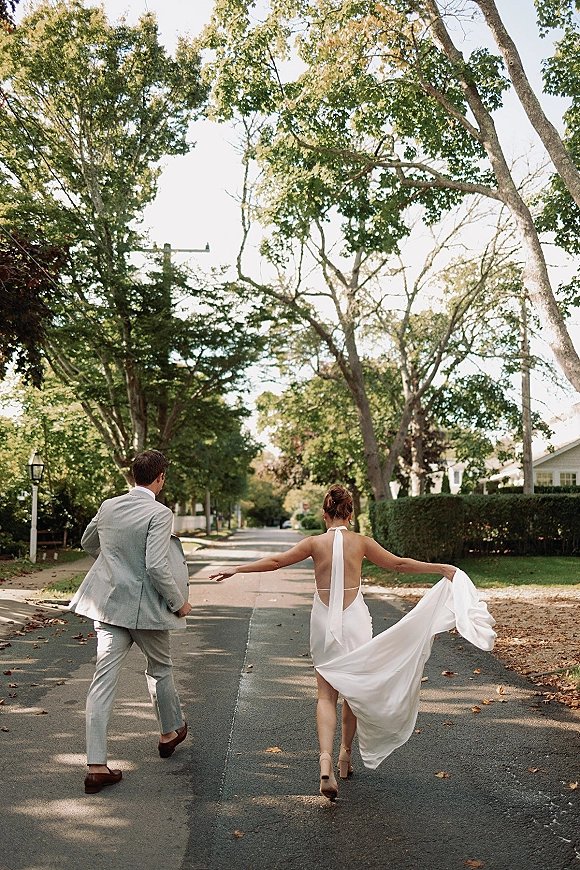 Couple portrait of bride and groom walking away, her open-back halter dress train in hand, on a tree-lined neighborhood street with leaves