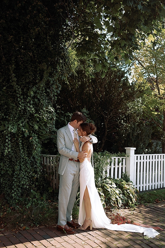 Couple portrait of bride and groom embrace, sharing a forehead kiss on a brick pathway beside an ivy wall and white picket fence