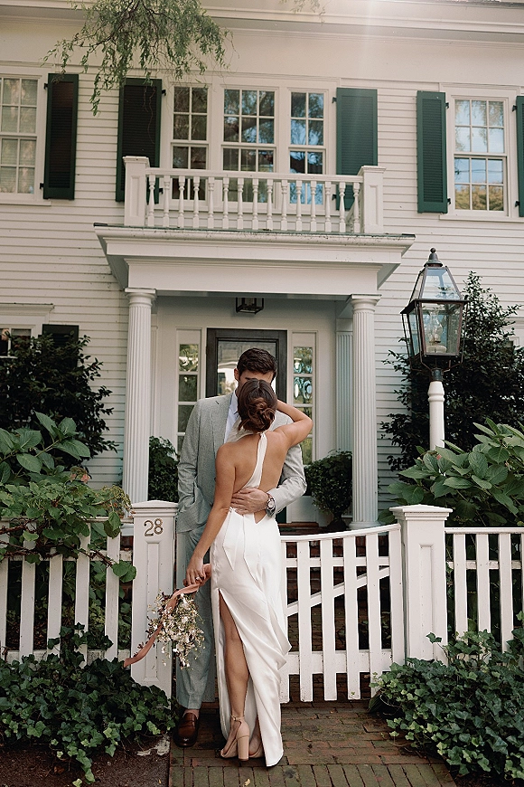 Couple portrait of bride and groom embrace, her open-back halter gown and bouquet with trailing ribbon by a white house porch and brick walkway