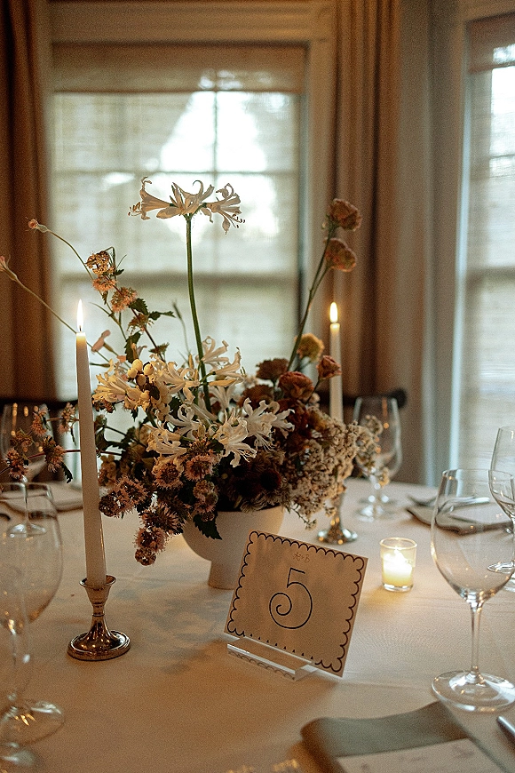 Reception tablescape with a wedding table centerpiece of wildflowers, taper candles, and wine glasses on a white tablecloth by curtained windows