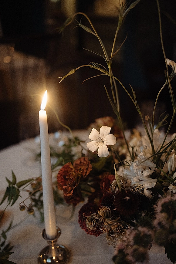 Wedding centerpiece with a taper candle centerpiece in a silver holder, surrounded by wildflowers and greenery on a white tablecloth in a dim reception
