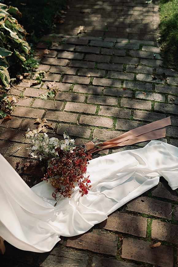 Bridal bouquet resting on a wedding dress train with a blush ribbon, set on a brick walkway amid garden foliage and dappled sunlight