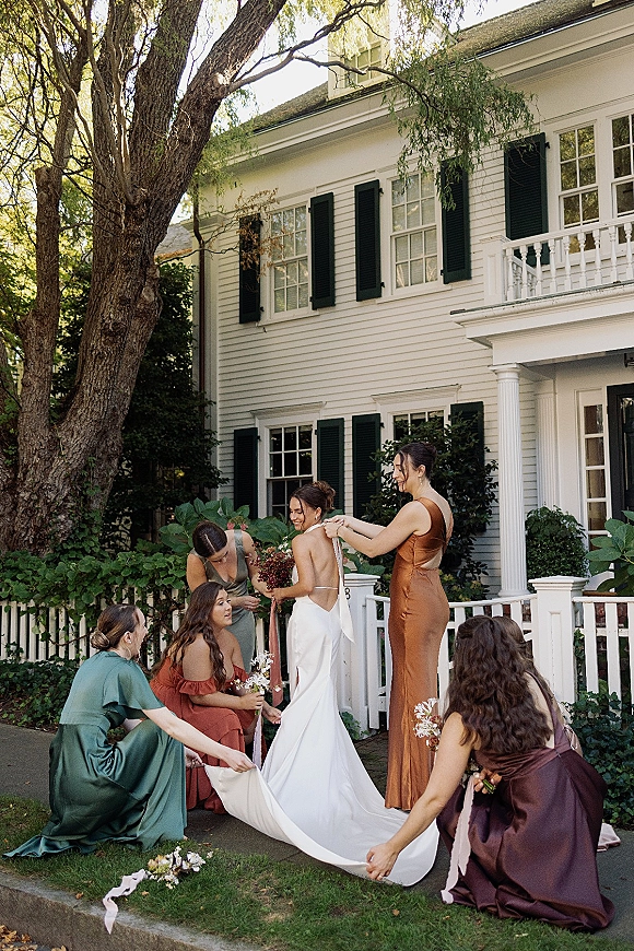 Bride with bridesmaids, bridal party getting ready as they adjust her dress train and hold bouquets on a white house porch with shutters