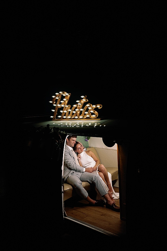 Couple portrait of bride and groom sitting in a car at night, bride in strapless dress leaning on groom in light gray suit, string lights and marquee sign glowing