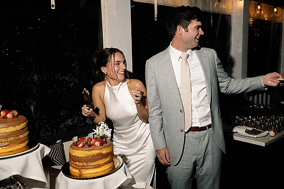 Wedding cake cutting as bride in halter dress and groom in light gray suit slice a berry-topped cake under patio string lights at night