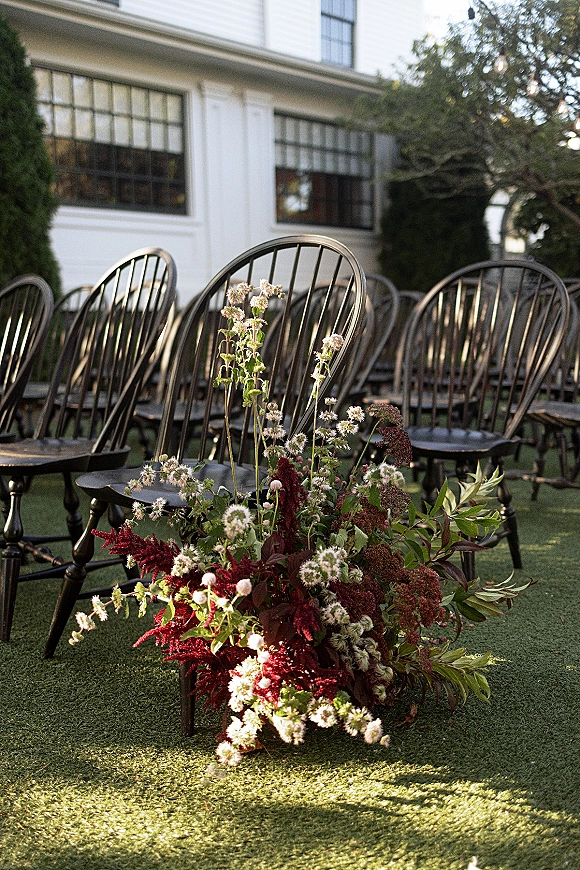 Ceremony aisle decor with wedding aisle flowers and low greenery arrangements beside black wooden chairs on a lawn under string lights