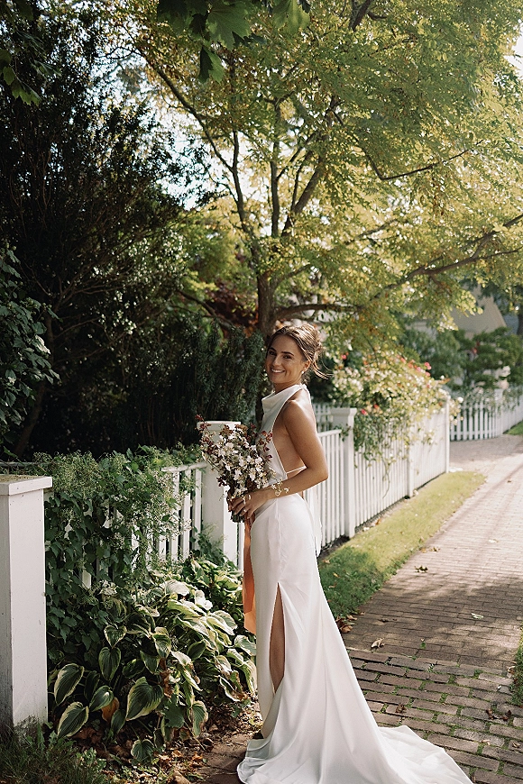 Bridal portrait of a smiling bride holding a bouquet, looking over her shoulder in a backless gown on a brick path by a white picket fence