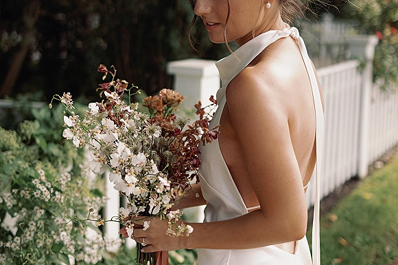 Bridal portrait of a bride holding bouquet in a white halter backless wedding dress, pearl drop earrings, by a white picket fence in a garden