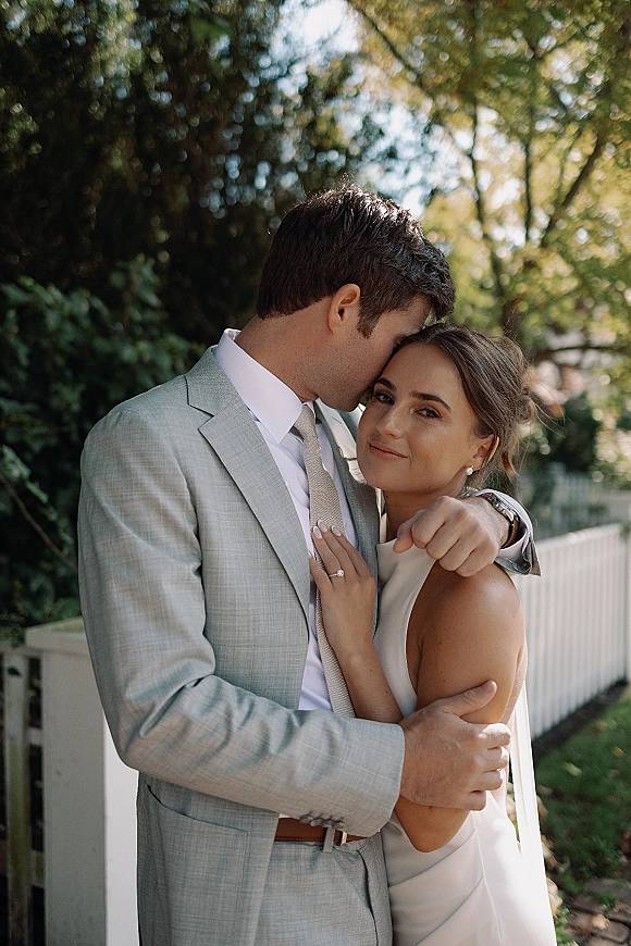 Couple portrait of bride and groom embrace as he kisses her forehead, showing pearl earrings and ring, with sunlit trees and fence behind