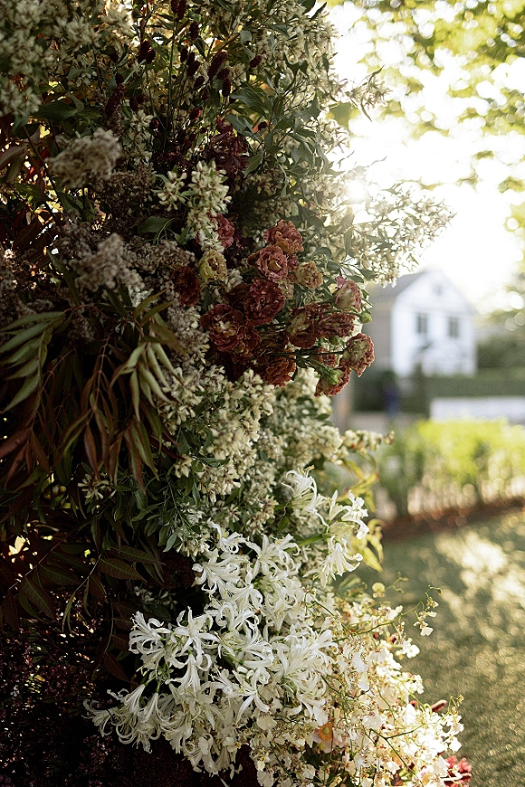 Wedding floral installation with a ceremony floral arch of white and burgundy blooms and lush greenery in a sunlit garden lawn near a house