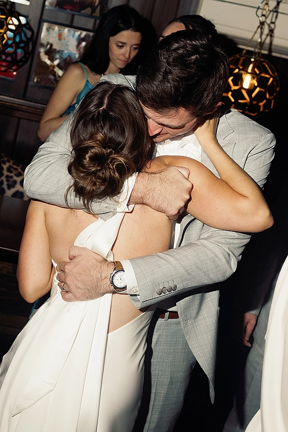 First dance moment as newlyweds sway close, bride in an open-back gown embracing groom in a suit under pendant lights with guests behind