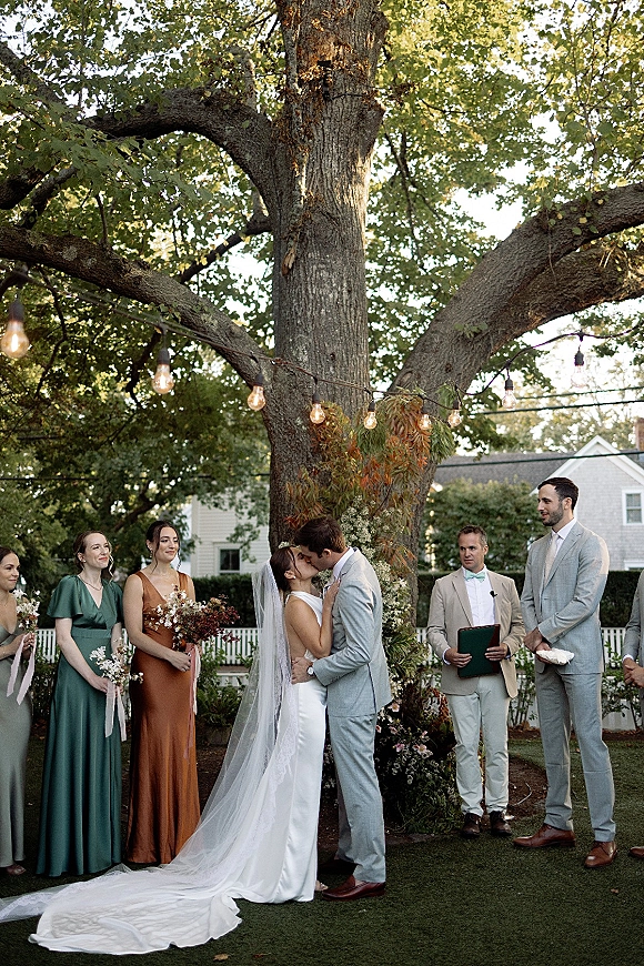 Ceremony kiss as bride and groom embrace beneath a greenery arch, veil trailing, wedding party nearby in a backyard garden setting