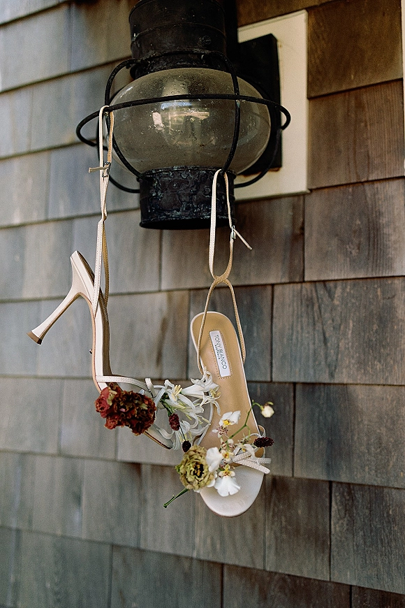 Wedding shoes hanging as white strappy heels with floral adornments, draped from a metal wall lantern against shingle siding