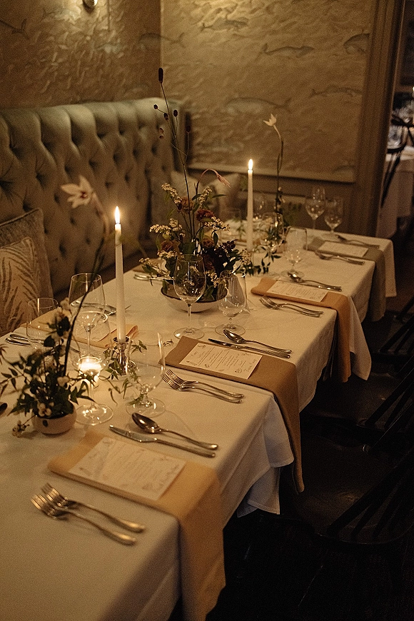 Reception tablescape with candlelit wedding table details, taper candles and floral greenery centerpiece on white linens in a dim restaurant setting