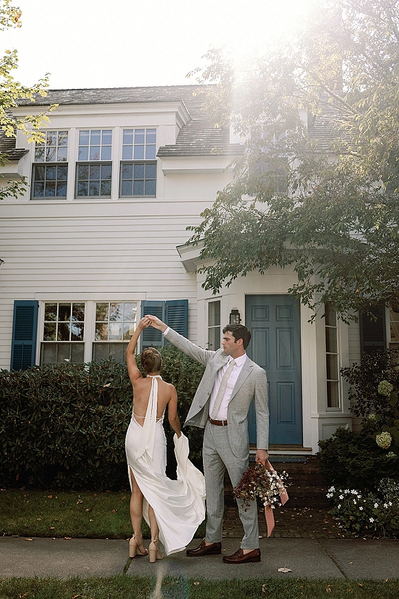Couple portrait of bride twirling pose beside groom holding bouquet, on front steps by a blue front door and white house shutters