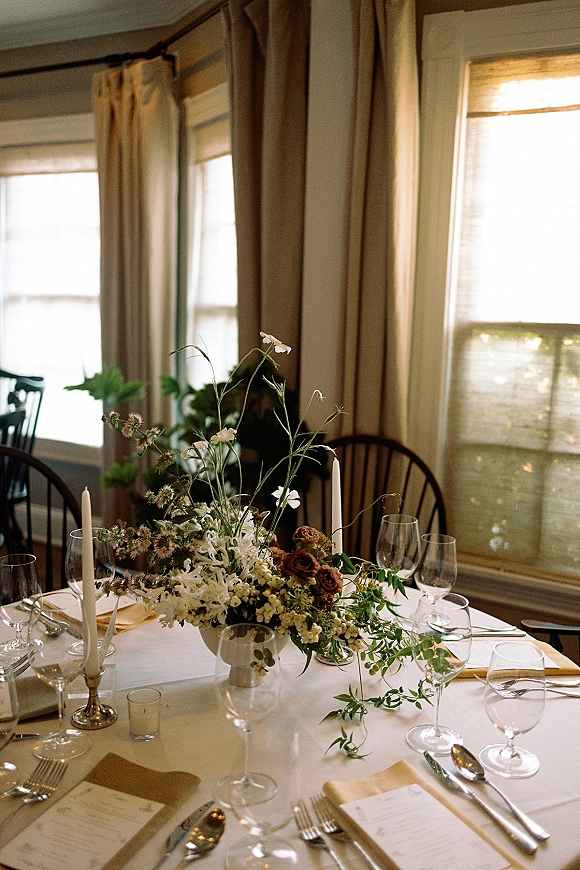 Reception tablescape with wedding table centerpiece, greenery, and white taper candles on a linen table set by curtained windows in a dining room