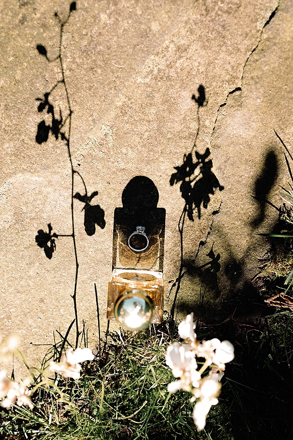 Engagement ring diamond solitaire in an open ring box beside a perfume bottle, styled on sunlit stone with floral shadows