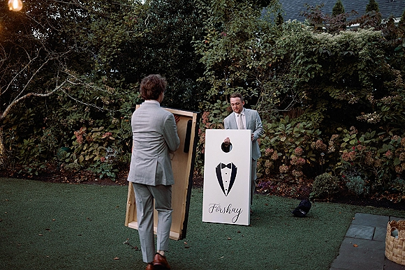 Wedding lawn games with groomsmen playing wedding cornhole on tuxedo boards, in light gray suits on a garden lawn under string lights